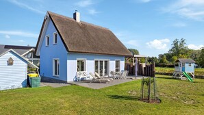 A blue house with a thatched roof, a fenced patio area with lounge chairs, and a small play structure in the background. - Nice home in Glowe with WiFi (Glowe)