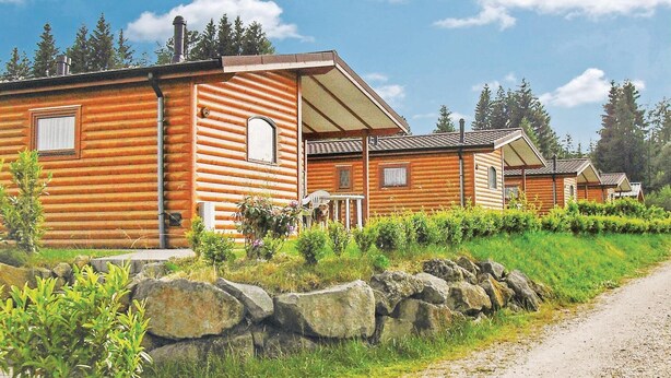 Une rangée de chalets en rondins, avec un mur de pierres en façade, un chemin de gravier sur la droite et un ciel bleu dégagé.