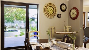 Uma sala de jantar com uma mesa posta para dois, vista do quintal através de uma porta de vidro deslizante e três relógios na parede.