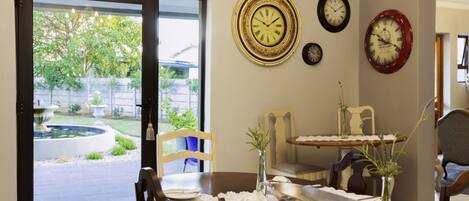 A dining area with a table set for two, a view of the backyard through a sliding glass door, and three clocks on the wall.