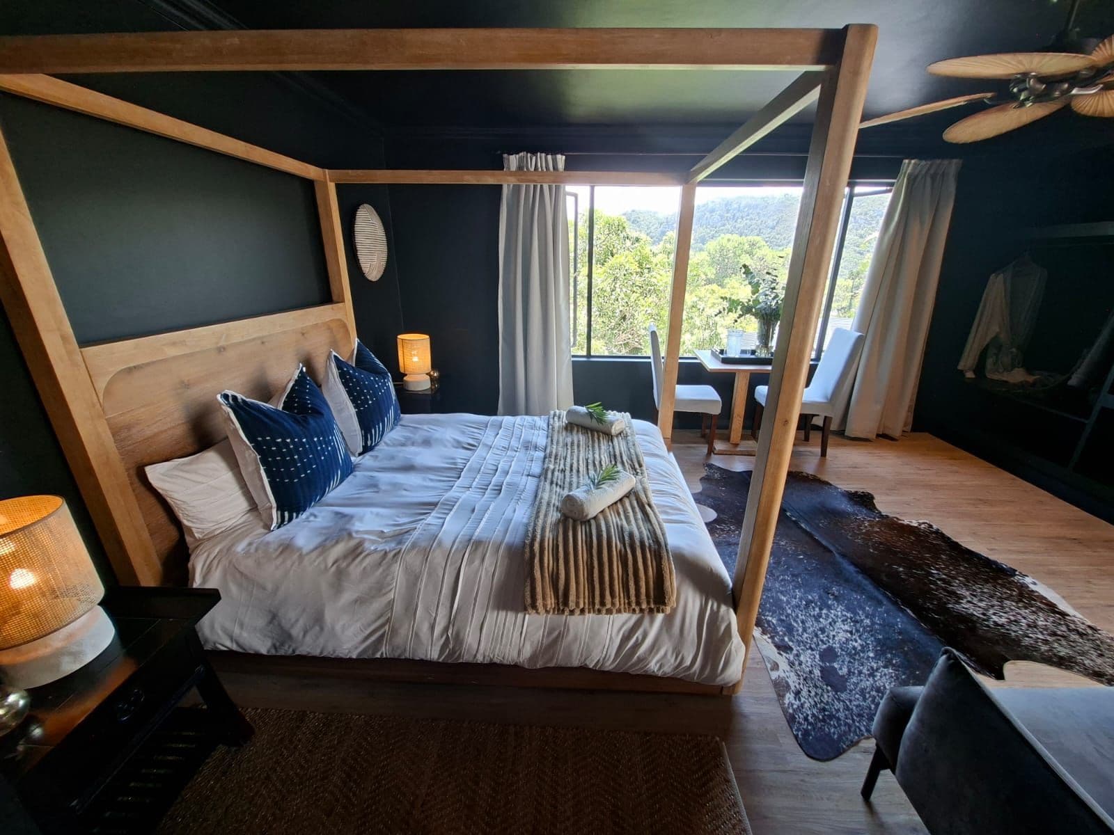 A four-poster bed with a wooden headboard, a bedside table with a lamp, and a view of the outdoors through a window.
