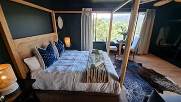 A four-poster bed with a wooden headboard, a bedside table with a lamp, and a view of the outdoors through a window.