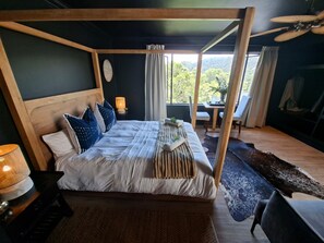 A four-poster bed with a wooden headboard, a bedside table with a lamp, and a view of the outdoors through a window. - Forest Nature Spa (Plettenberg Bay)