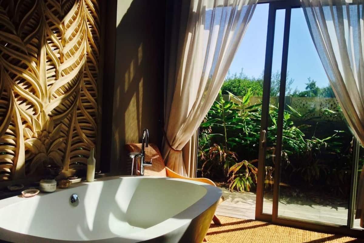 A modern bathroom with a freestanding bathtub, a decorative wall panel, and a view of lush greenery through large sliding glass doors.