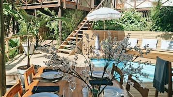 A wooden dining table set for a meal with a view of a pool and a staircase leading to an upper level.