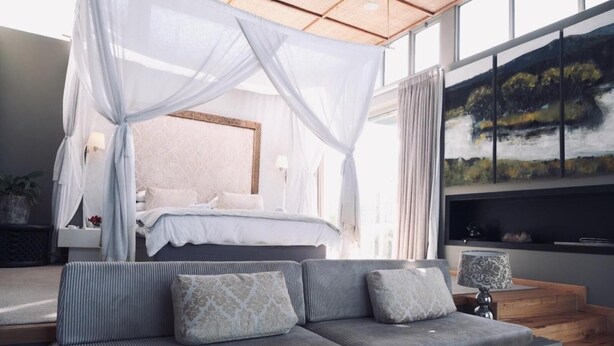 Ein Schlafzimmer mit Baldachinbett, grauem Sofa mit gemusterten Kissen, Nachttisch mit Lampe und einem Gemälde an der Wand.