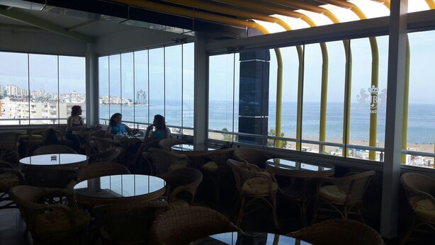 An indoor seating area with wicker chairs and tables, large windows offering a view of the sea and buildings. - Jasmin Blue Otel (Silifke)