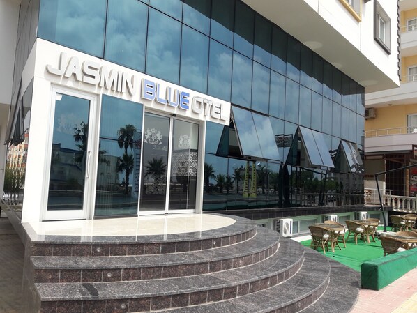 The exterior of Jasmin Blue Hotel with a modern glass facade, a set of stairs leading to the entrance, and outdoor seating area.
