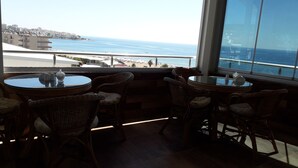 A balcony with wicker chairs and a glass table, overlooking a coastal view with buildings and a beach. - Jasmin Blue Otel (Silifke)