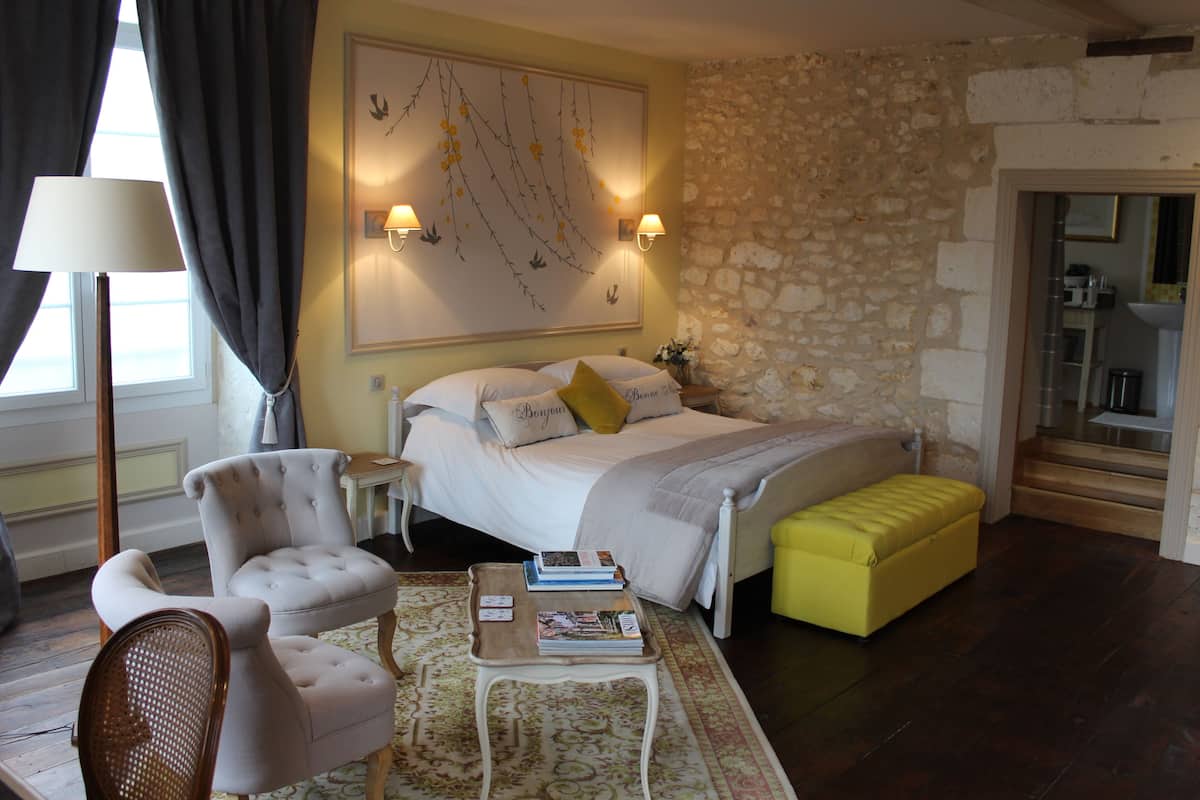 Een slaapkamer met een bed, een nachtkastje, een stoel, een bankje en een kunstwerk aan de muur.