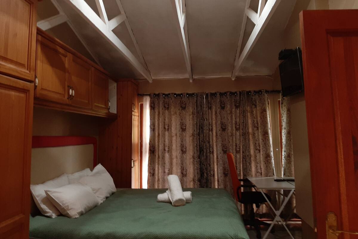 Een kamer met een bed, een bureau en een stoel. Er is een raam met gordijnen en een deur, en een handdoek op het bed.