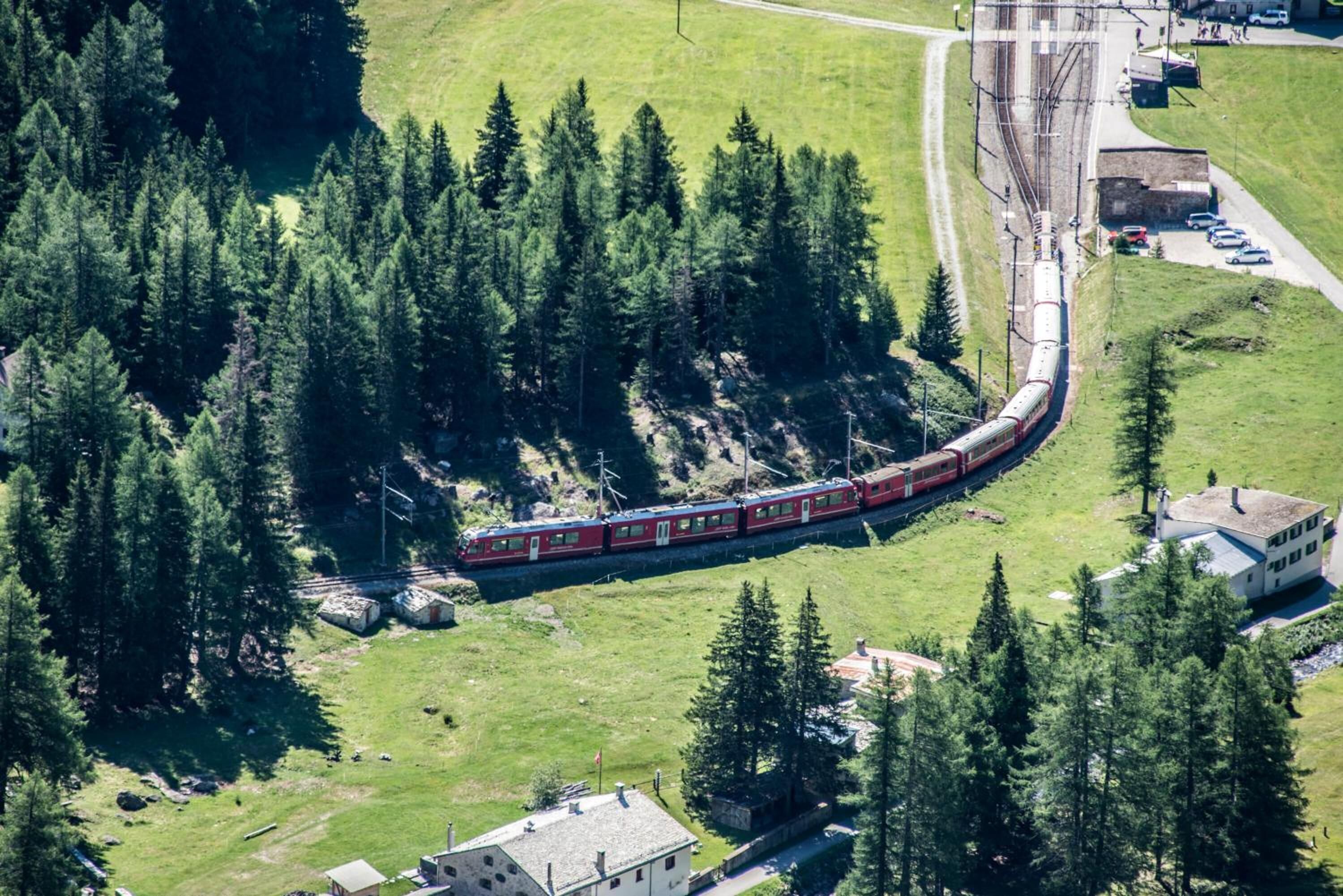 Un tren serpentea por una zona boscosa con un edificio pequeño y una carretera cerca.