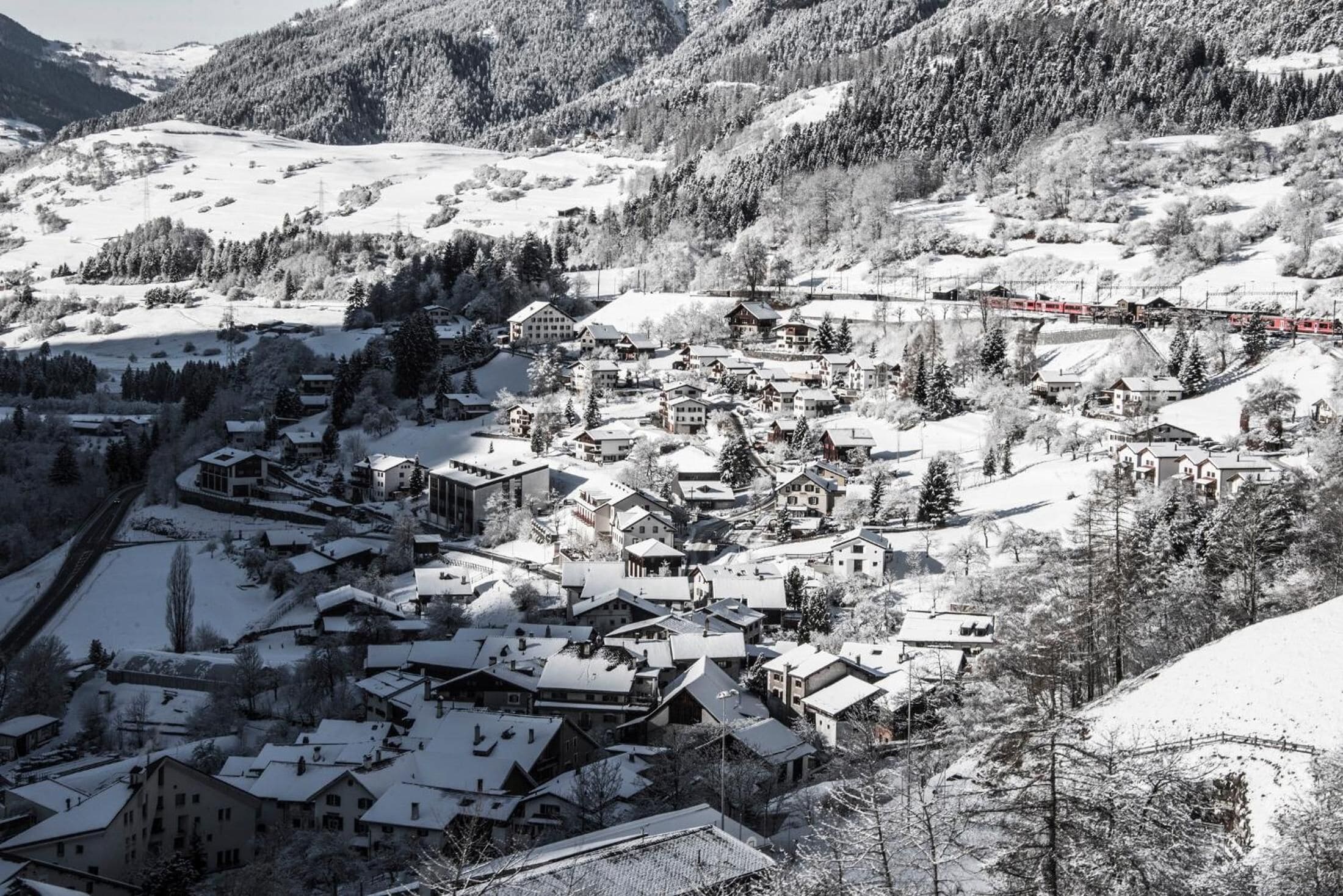 Uma aldeia coberta de neve com edifícios tradicionais, um trem e montanhas ao fundo.