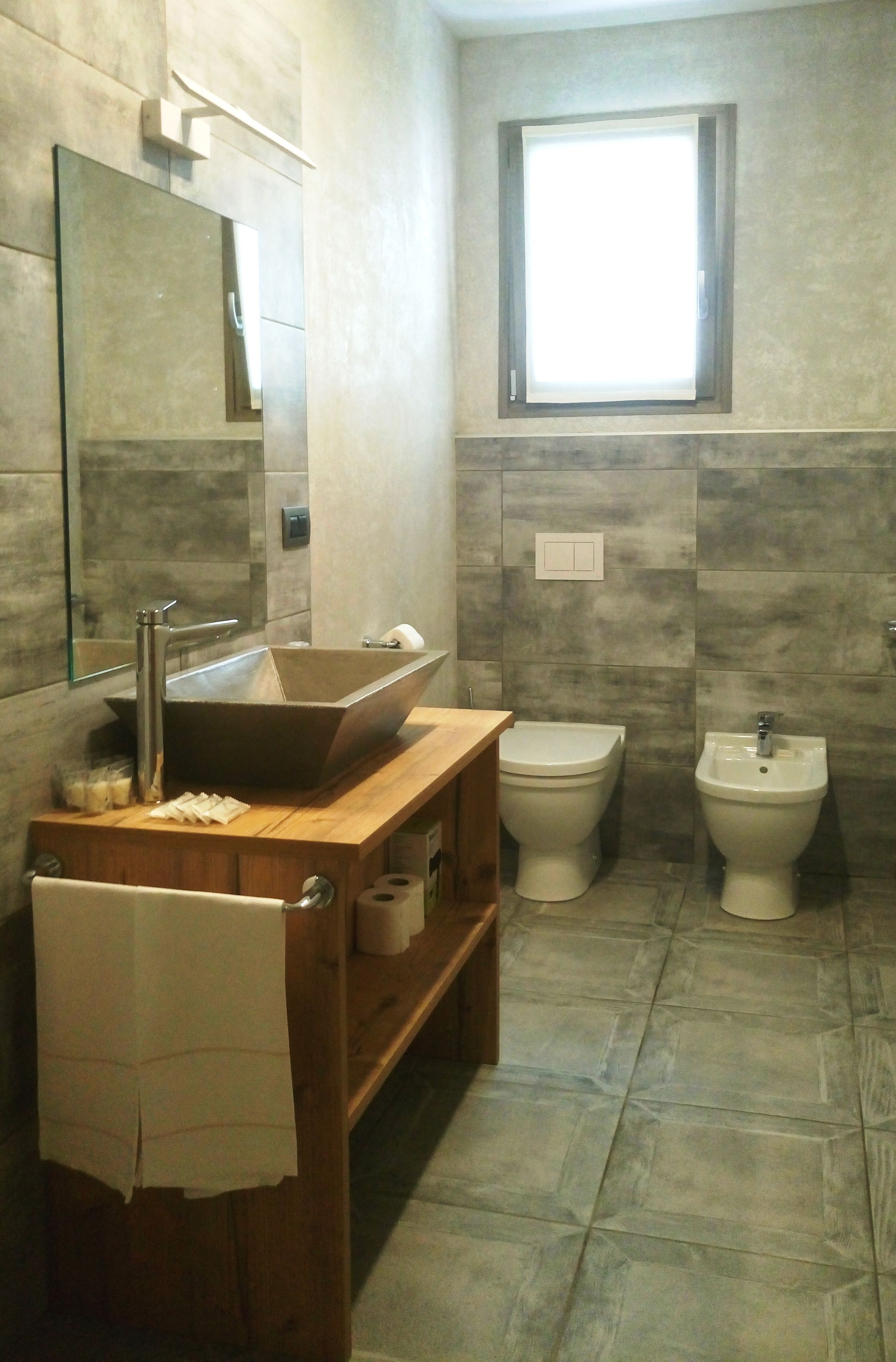 A modern bathroom with a wooden vanity, a large mirror, a vessel sink, two toilets, and a window.