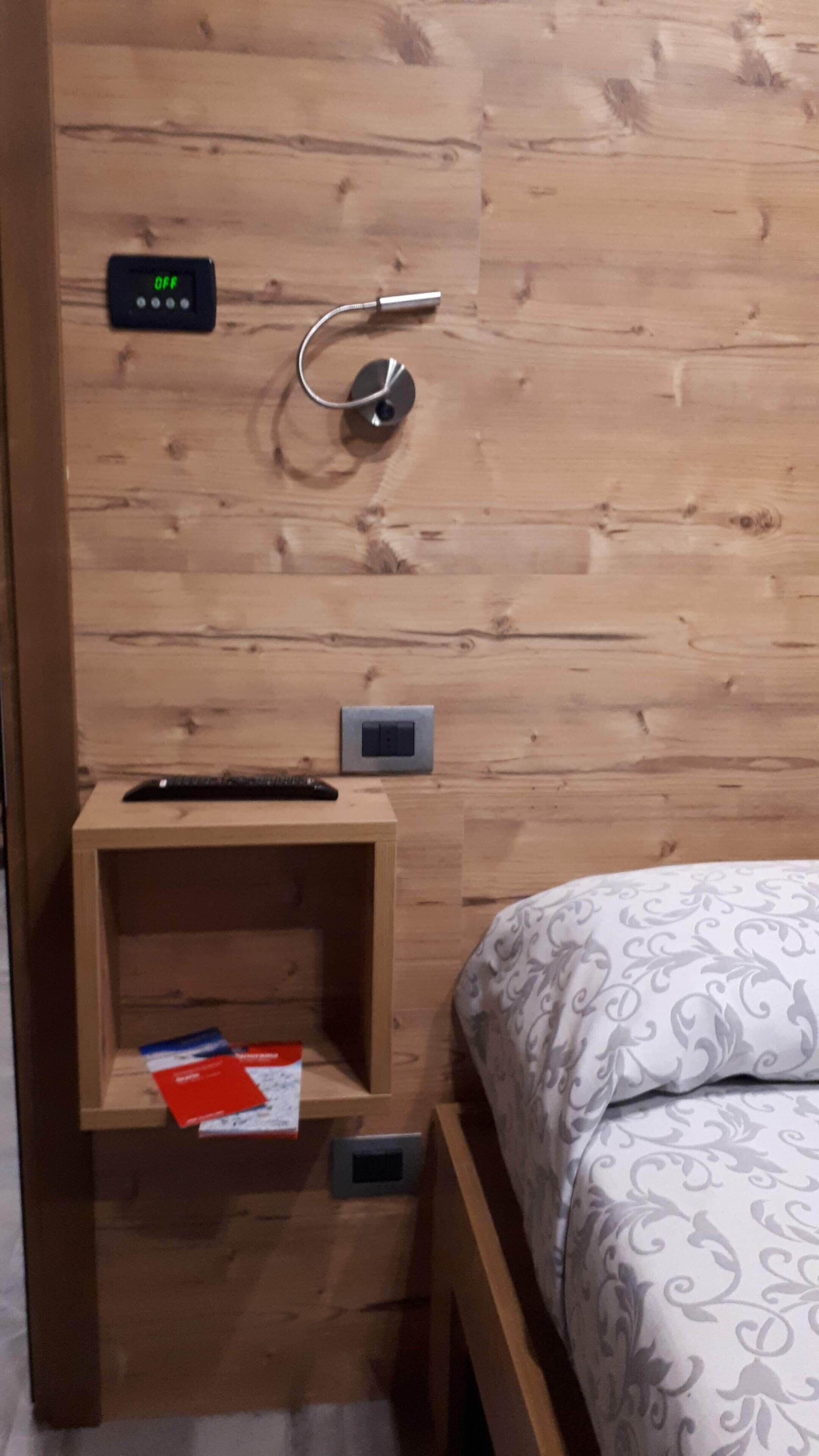 A wooden wall with a power outlet, a thermostat, and a remote control on a small wooden nightstand.