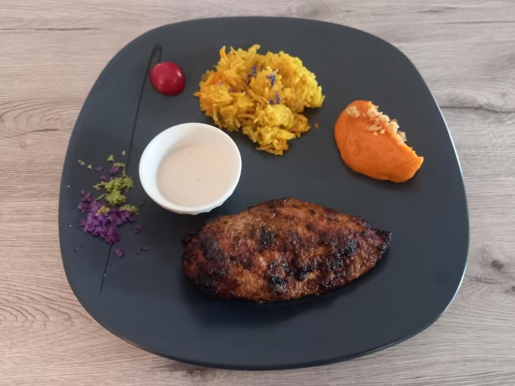 A plate with a grilled meat, mashed potatoes, a slice of sweet potato, a cherry, and a small bowl of sauce.