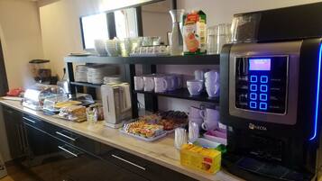 Un área de bufé con una máquina de café de autoservicio, diferentes opciones de comida y una selección de tazas y platos.