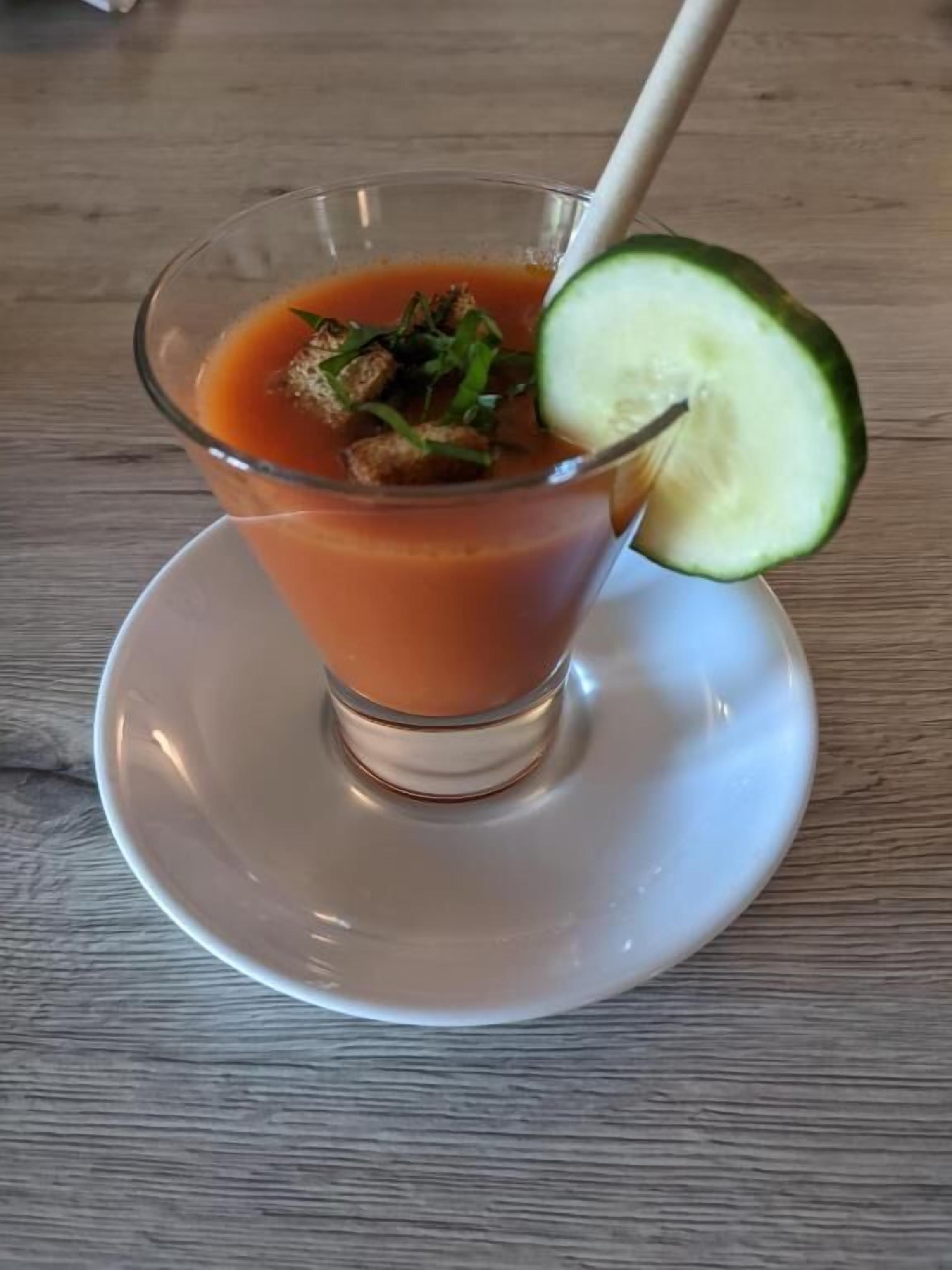 A glass of tomato-based drink with a cucumber garnish on a white plate.