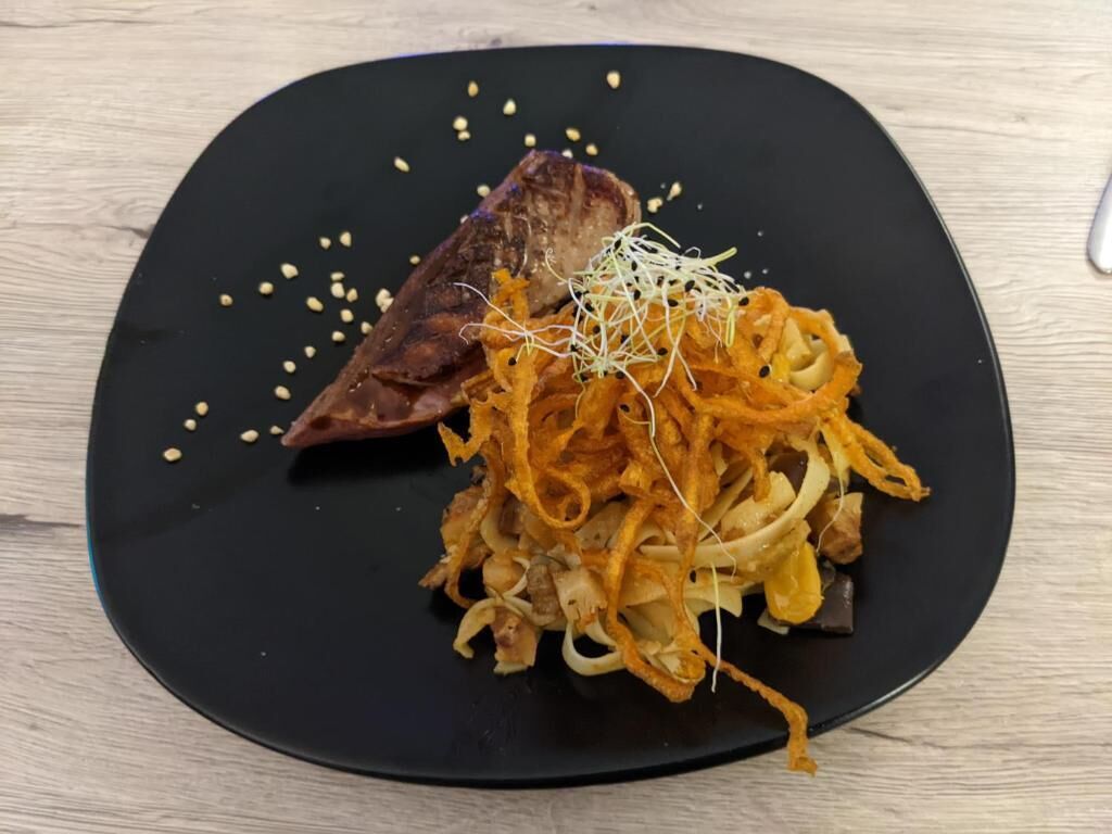 A black plate with a dish consisting of noodles, a piece of meat, and a garnish of sprouts and shredded carrots.