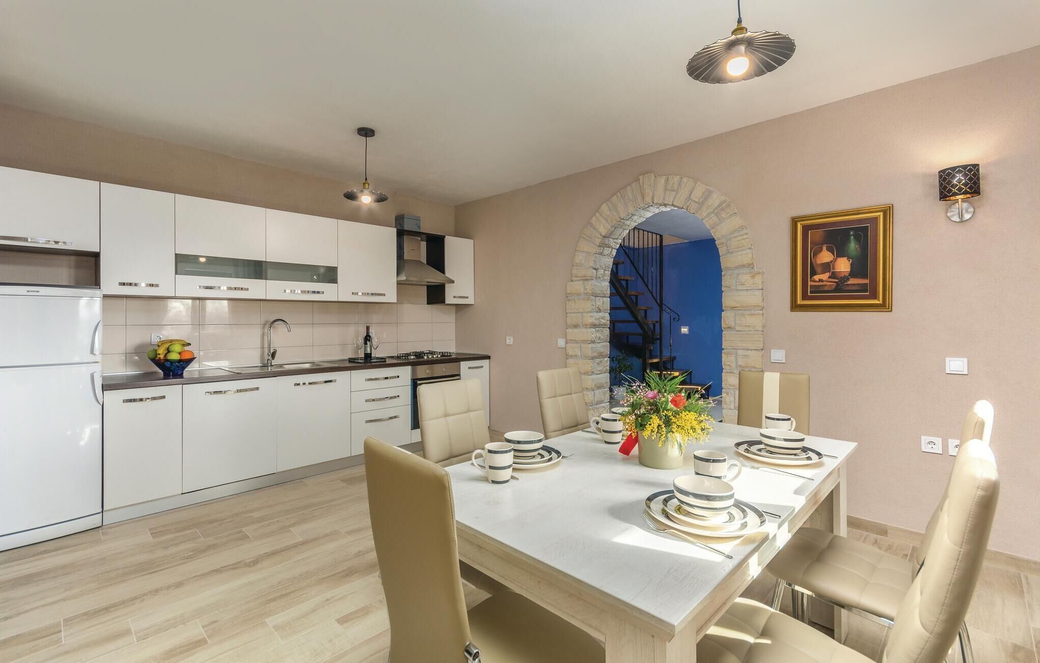 Une cuisine moderne avec des meubles blancs, un coin repas avec une table dressée pour quatre personnes, et un escalier bleu menant à l’étage supérieur.