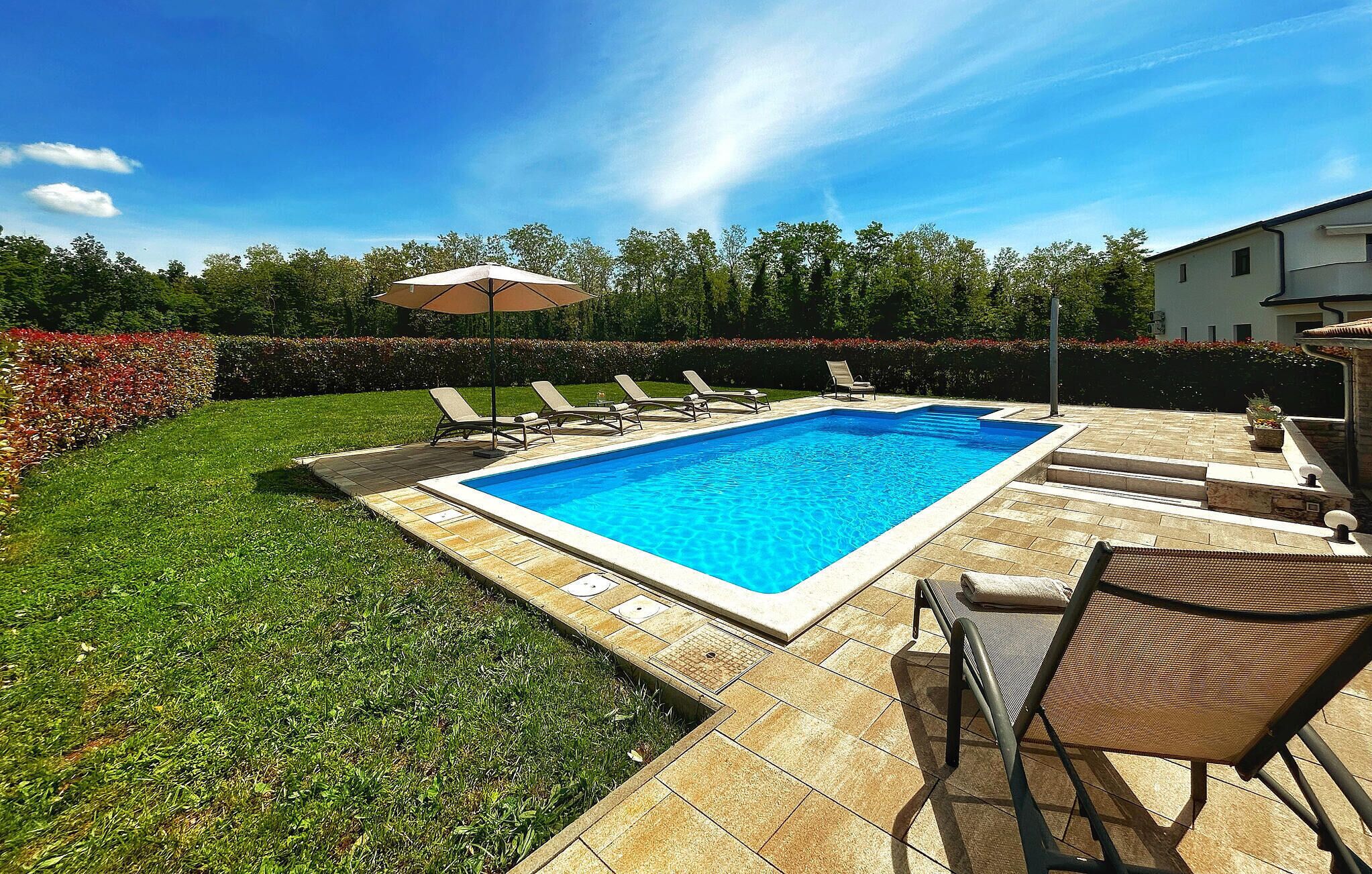 Una piscina rectangular con solárium empedrado, rodeada de césped y un seto.
