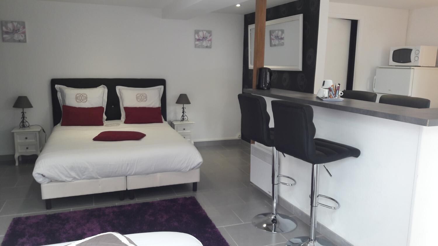 A modern hotel room with a bed, two bedside tables, a nightstand, a purple rug, a bar area with two stools, and a microwave on the counter.
