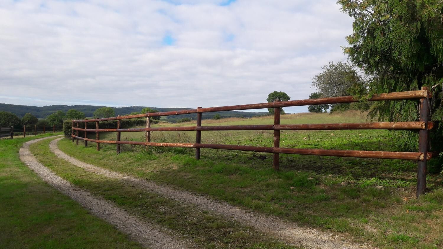 A rural road curving through a grassy field with a wooden fence on the right.