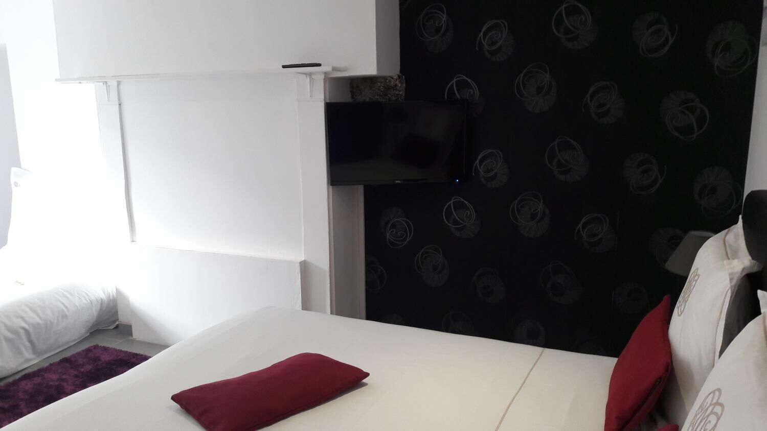 A hotel room with a white bed, a red pillow, a black wall with a circular pattern, and a mounted TV.
