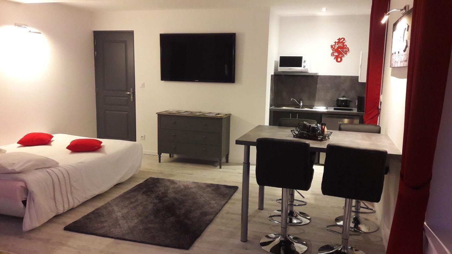 A compact hotel room with a bed, a desk with two chairs, a TV, a kitchenette, and a red decorative wall piece.