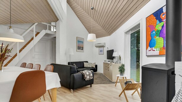 Un salon moderne avec un canapé noir, une table à manger en bois et des chaises. On y trouve un escalier menant à un autre niveau, un téléviseur et une grande fenêtre ornée d’une fresque colorée.