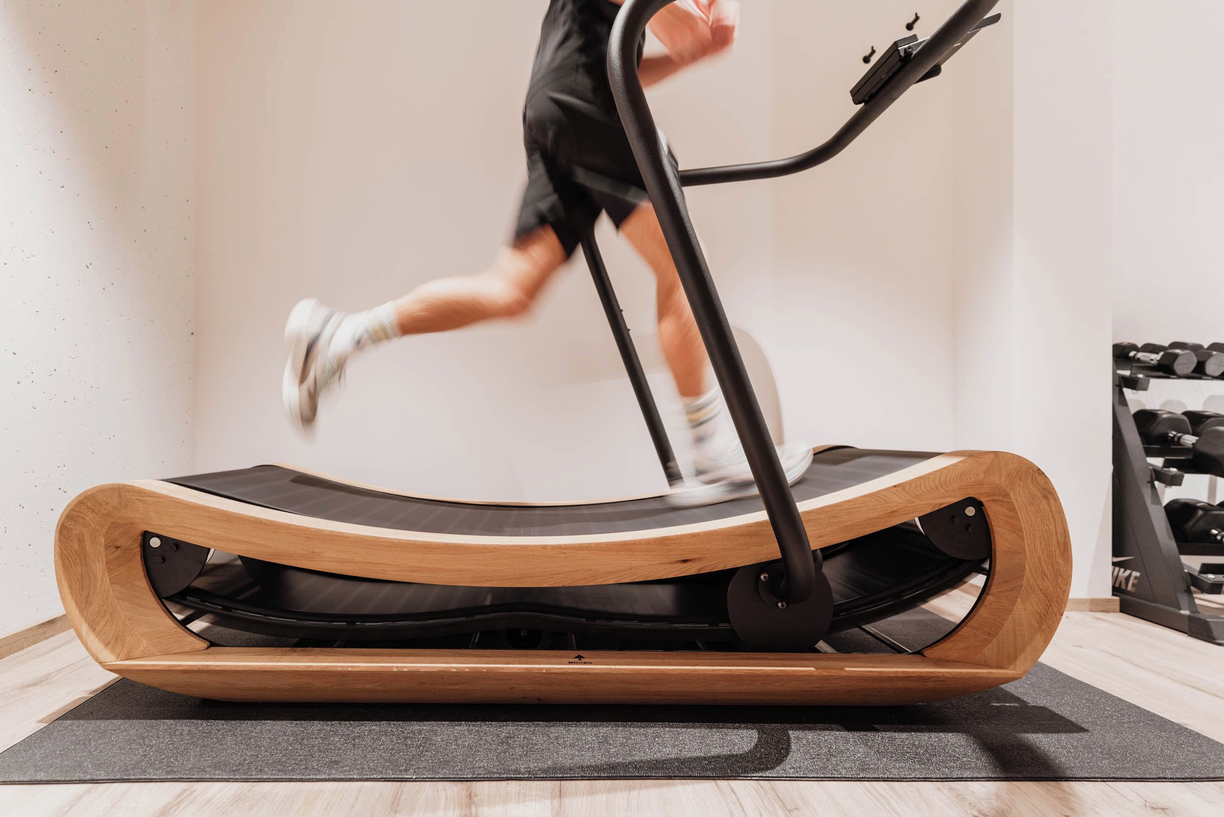 A person running on a treadmill in a room with a wooden floor and a wall-mounted dumbbell rack.