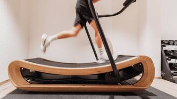 A person running on a treadmill in a room with a wooden floor and a wall-mounted dumbbell rack.