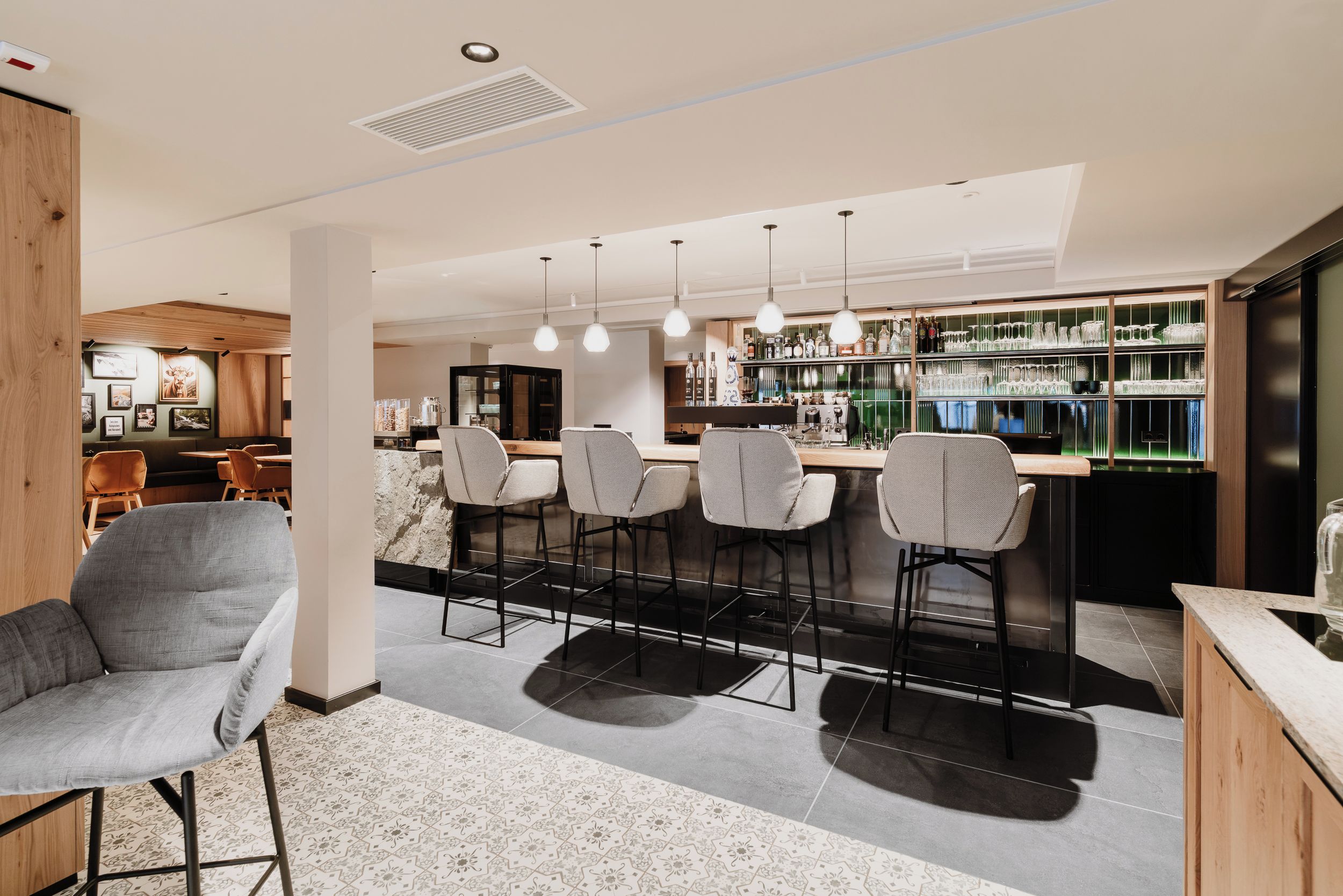 Un área de bar moderna con taburetes, iluminación colgante y una barra repleta de botellas y copas.