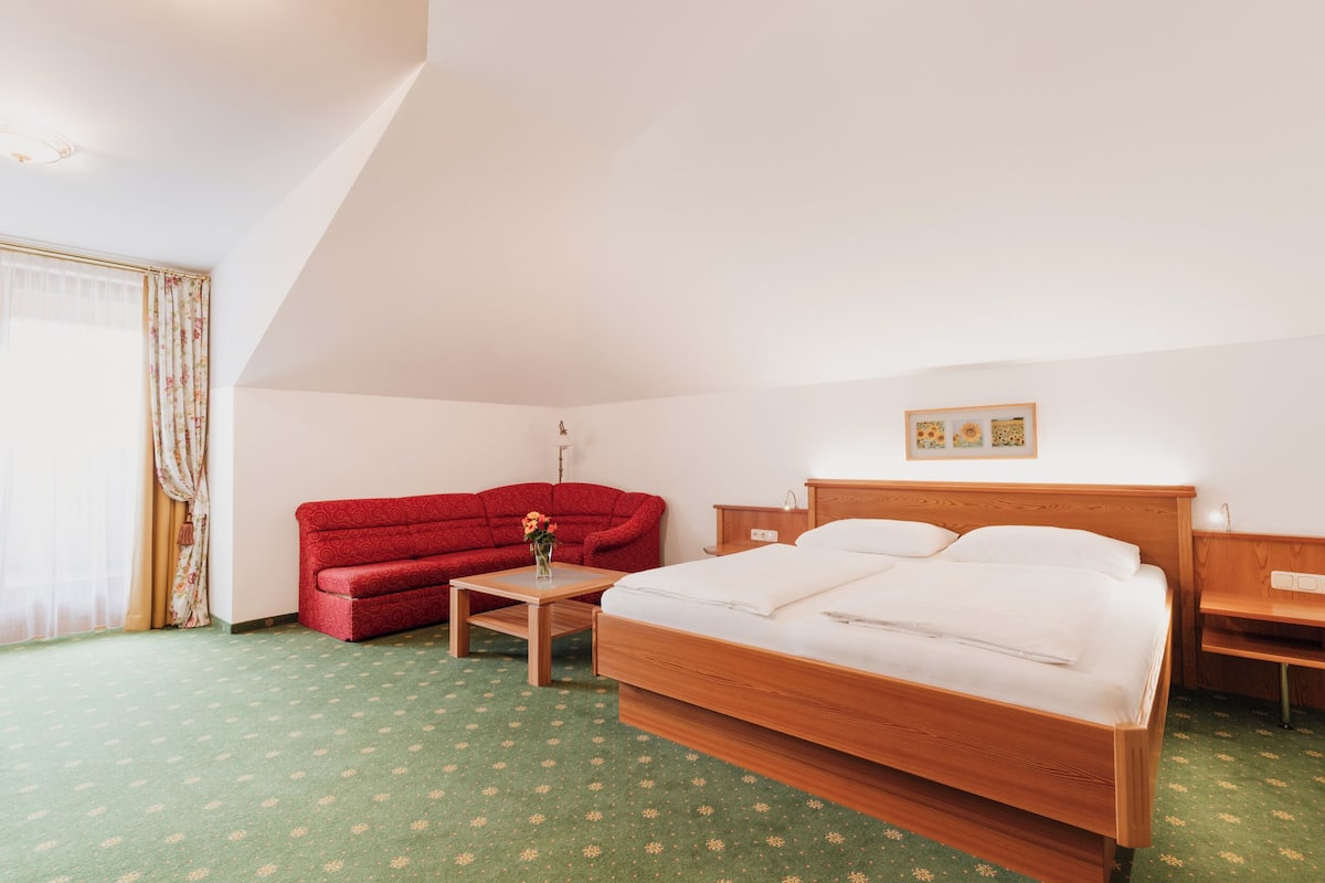 Una habitación de hotel con una cama, un sofá rojo, una mesita con un jarrón y una mesita de noche de madera.