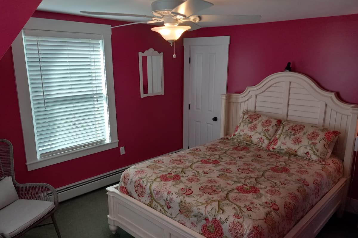 A bedroom with a bed, a chair, a window with blinds, a door, and a ceiling fan.