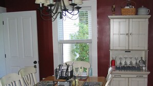 A dining room with a wooden table, white chairs, a chandelier, and a kitchen area with cabinets and a window. - Bittersweet Bed and Breakfast (York Beach)