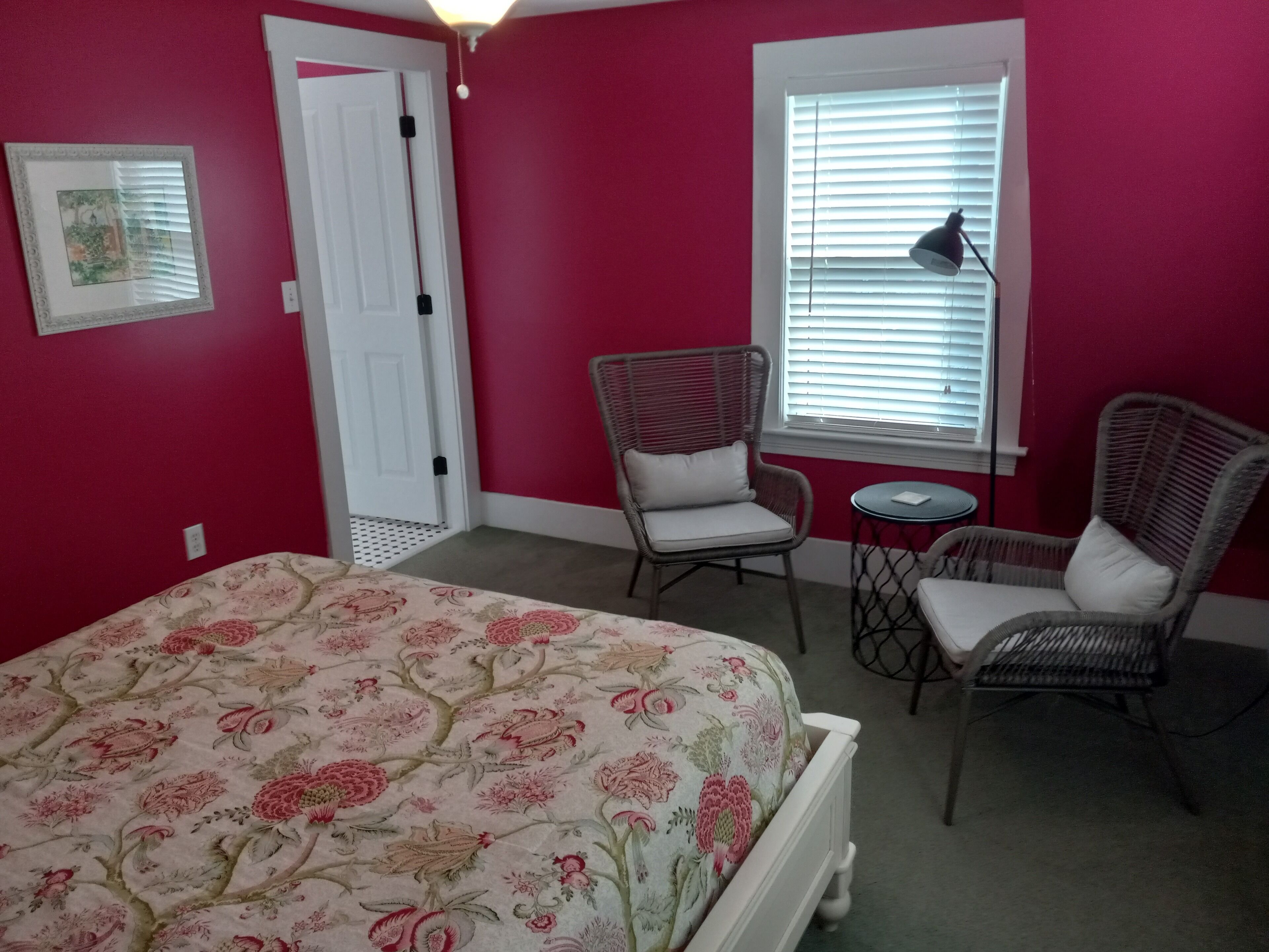 A bedroom with a floral bedspread, two wicker chairs, a small round table, and a window with blinds.