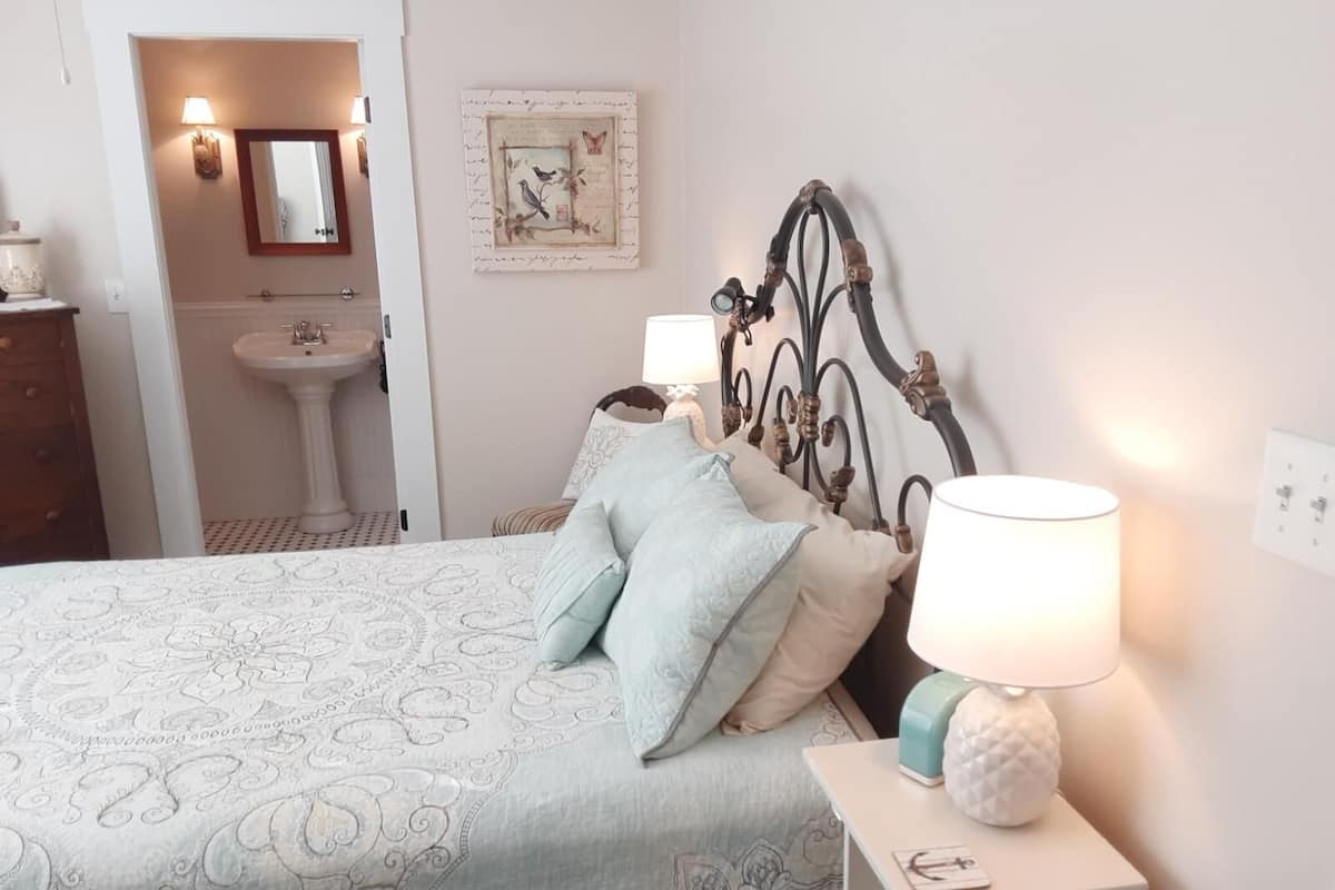A bedroom with a bed, bedside lamps, a nightstand, a wall-mounted mirror, a decorative piece, and a bathroom with a sink.