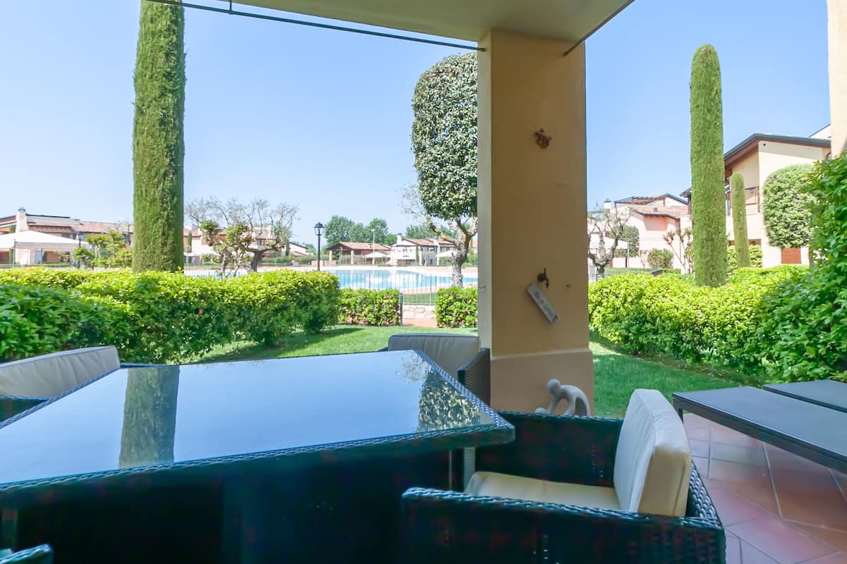 Une terrasse avec une table en verre et des chaises en osier, une piscine en arrière-plan et des haies soigneusement taillées.