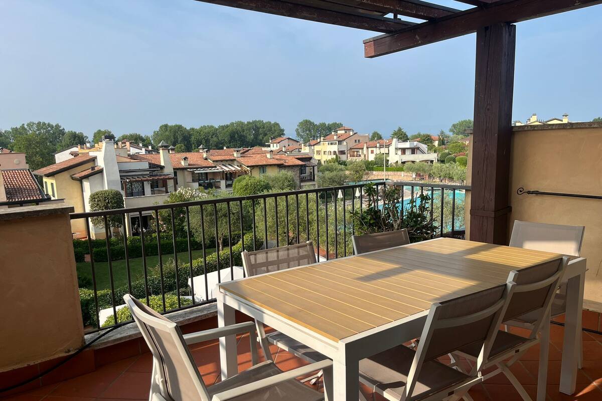 Un balcon avec une table et des chaises en bois, une piscine en arrière-plan et une vue sur les maisons environnantes.