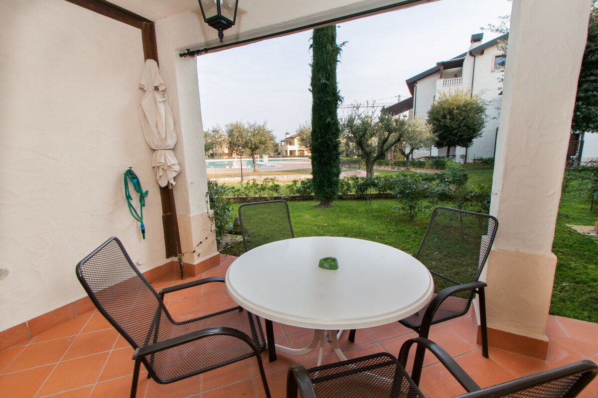 Une terrasse avec une table ronde et quatre chaises, un parasol blanc, et une vue sur une piscine et la verdure environnante.