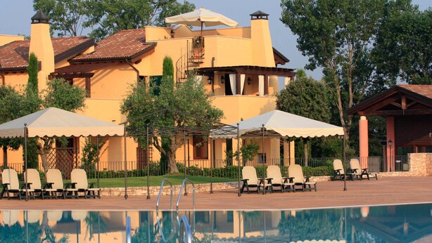 A pool area with lounge chairs and umbrellas, a building with a tiled roof, and a clear sky. - Lugana Village Resort & Sporting Club (Peschiera del Garda)