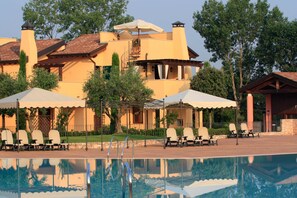 Un espace piscine avec des chaises longues et des parasols, un bâtiment doté d’un toit en tuiles et un ciel dégagé.