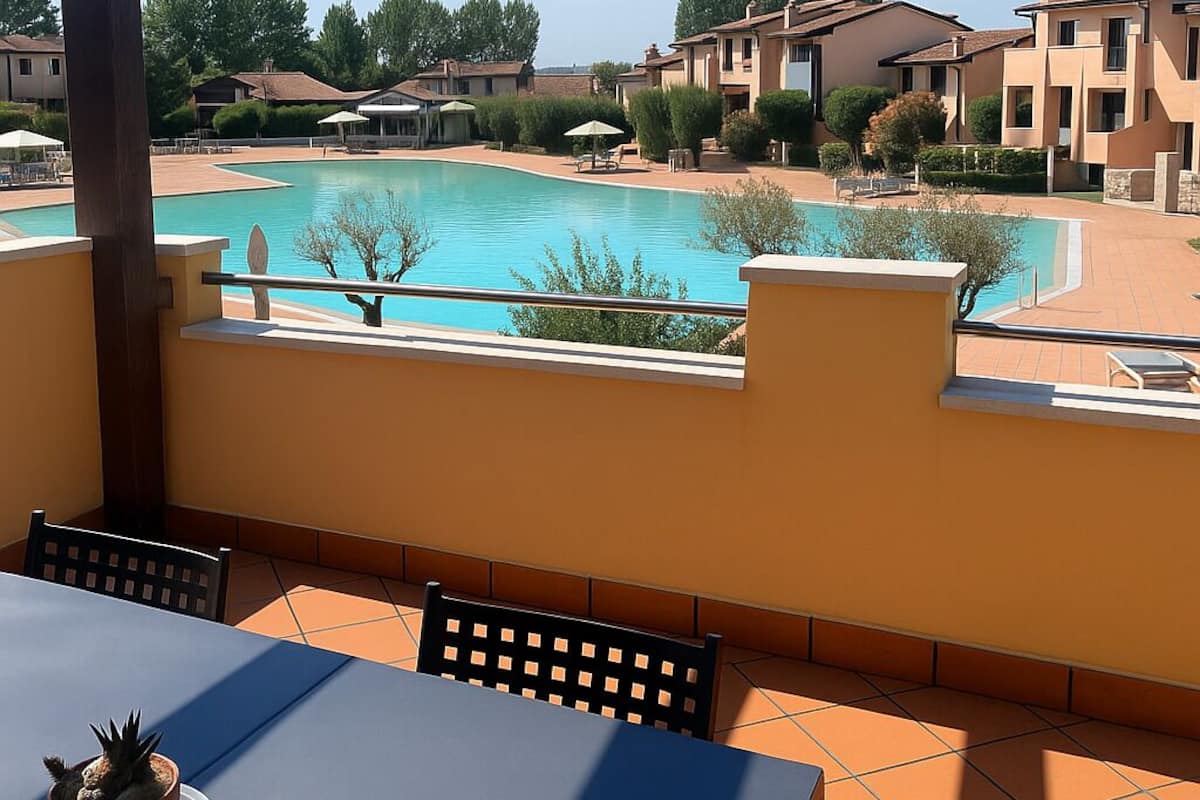 Une vue sur la piscine, avec une table et des chaises, une plante en pot, et une piscine d’eau cristalline entourée de bâtiments.