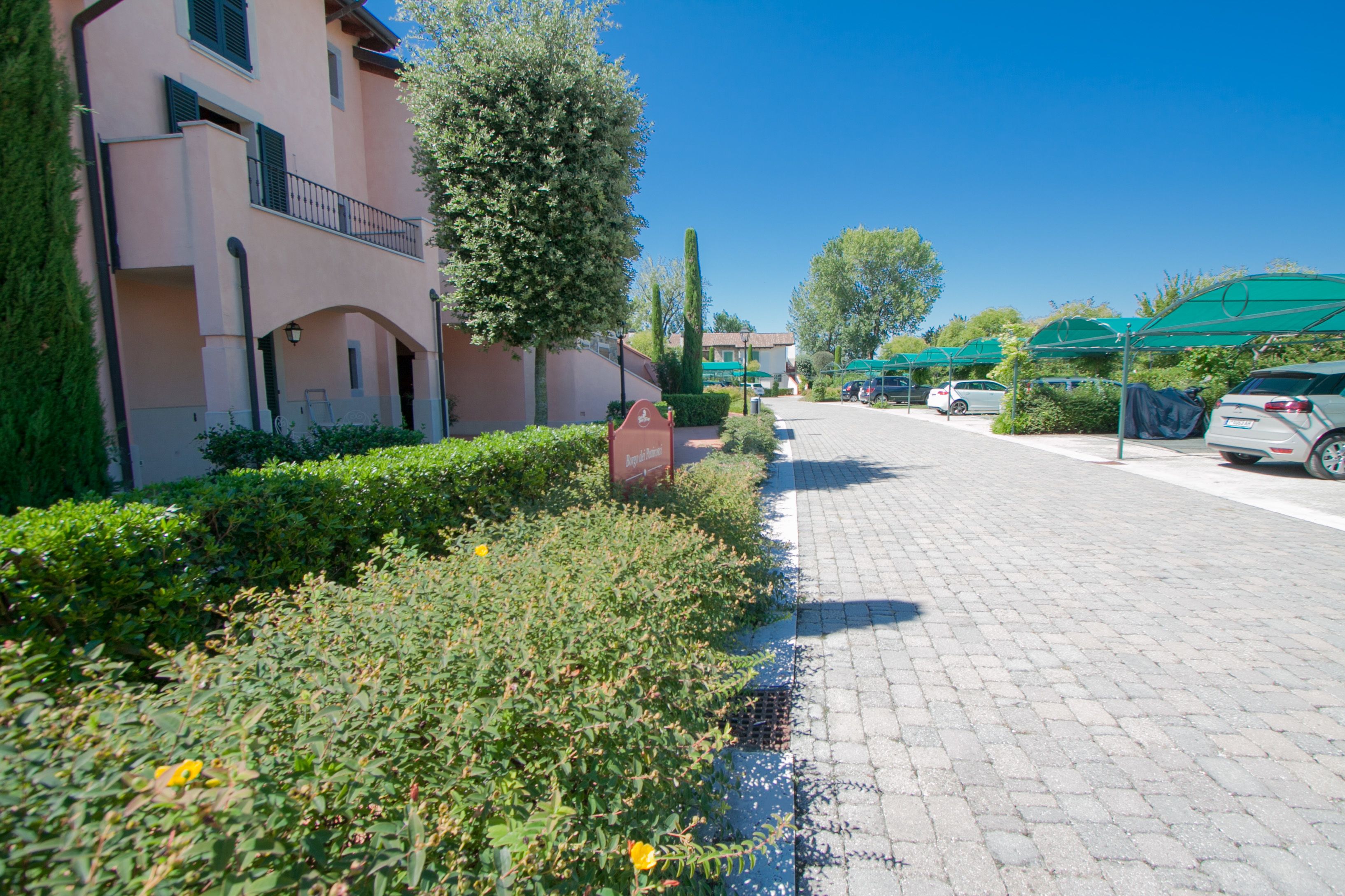 A paved driveway leading to a building with a balcony, surrounded by greenery and parked cars.