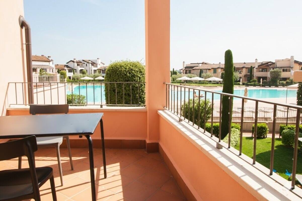 Un balcon avec une table et des chaises, donnant sur une piscine et des immeubles d’habitation.