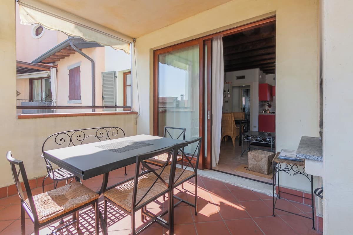 Un balcon avec une table et des chaises, une porte-fenêtre coulissante donnant sur une cuisine et une vue sur les immeubles voisins.