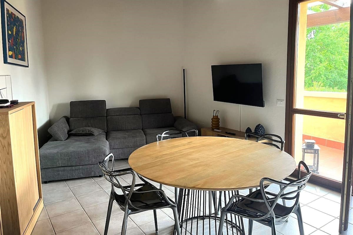 Un salon moderne avec un canapé d’angle gris, une table à manger en bois avec quatre chaises, un téléviseur à écran plat fixé au mur et une grande fenêtre donnant sur un paysage verdoyant.