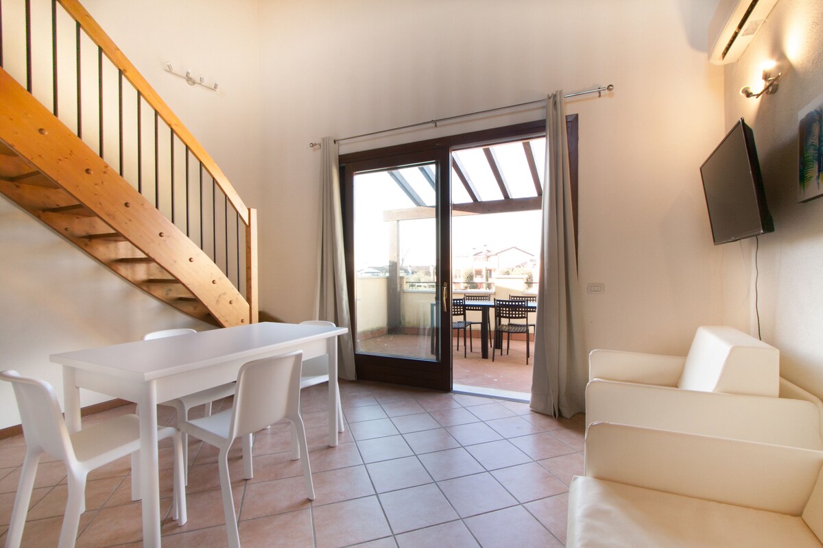 Un salon moderne avec un canapé blanc, une table à manger avec des chaises, un escalier en bois et une porte-fenêtre coulissante donnant sur un patio extérieur.