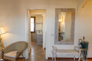 Une pièce comprenant un fauteuil en osier, un banc, un miroir avec un cadre orné et une porte donnant accès à une salle de bain.